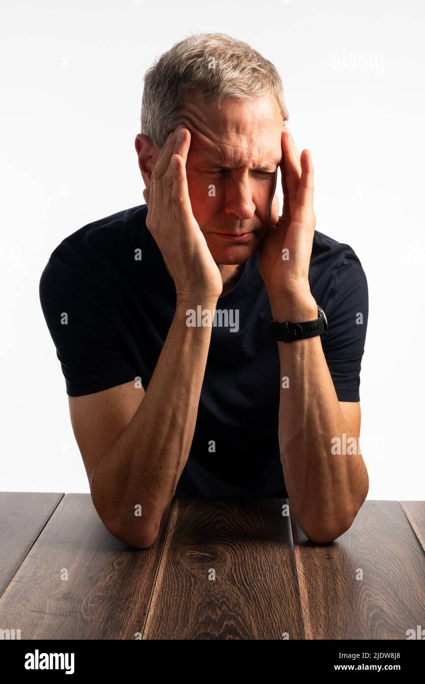 Älterer Mann in einem blauen T-Shirt, der seinen Kopf hält, auf einem dunklen Holztisch mit einem reinen, brillanten weißen Hintergrund, der schmerzt, gestresst und besorgt aussieht Stockfoto