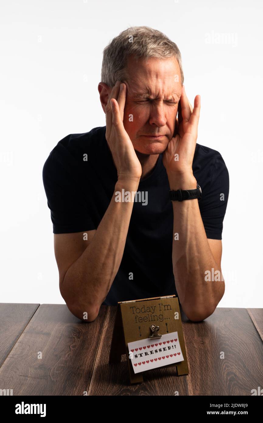 Älterer Mann in einem blauen T-Shirt, der seinen Kopf hält, auf einem dunklen Holztisch mit einem reinen, brillanten weißen Hintergrund, der schmerzt, gestresst und besorgt aussieht Stockfoto