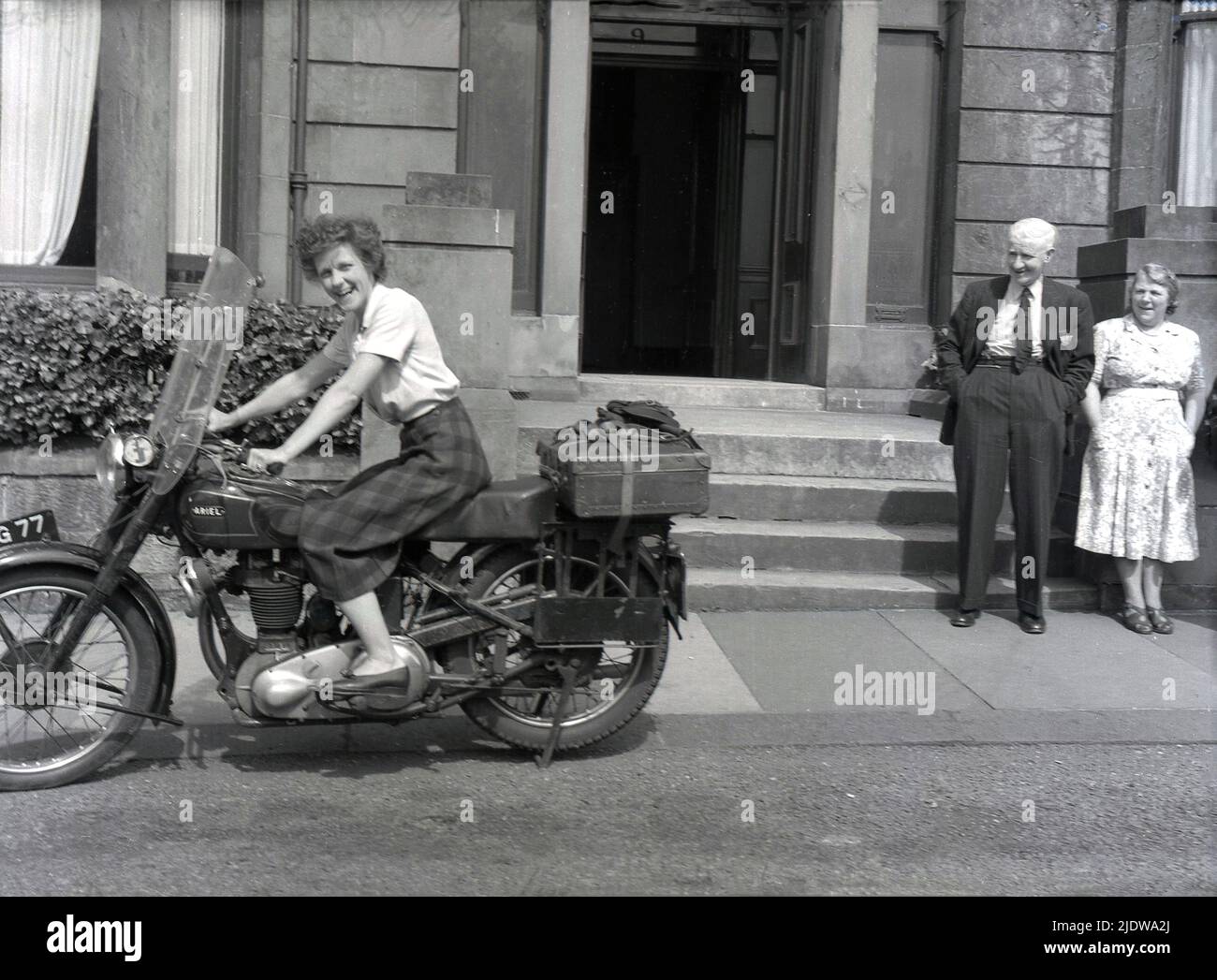 1956, historisch, eine Dame in einem Tartanrock, die auf einem Ariel-Motorrad der damaligen Zeit vor einem Hoteleingang in der Nähe von Mallaig, schottischen Highlands, Schottland, Großbritannien, sitzt. Das Motorrad ist ein Ariel 'Square Four', auch bekannt als der 'Squariel'. Entworfen von Edward Turner, der 1928 den ersten „Square Four“-Motor entwickelt hatte, und hergestellt vom Briitsh-Hersteller Ariel Motors Ltd. Aus Birmingham aus dem Jahr 1953, hatte er einen 1000cc 4-Zylinder-Motor - den größten jemals in Großbritannien produzierten - was ihn ideal für Motorradtouren machte. Als die Nachfrage zurückging, wurde die Produktion des Modells 1959 eingestellt. Stockfoto