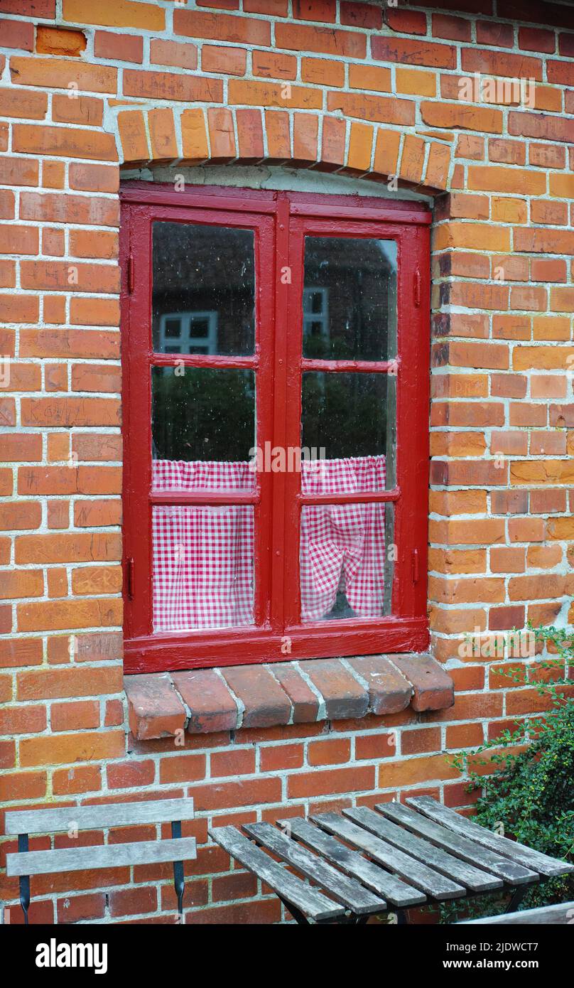 Altes Fenster eines Backsteinhauses oder eines Hauses. Altes rot gerahmtes Fensterflügel in einem historischen Gebäude mit einem kleinen karierten Vorhang. Äußere Details Stockfoto
