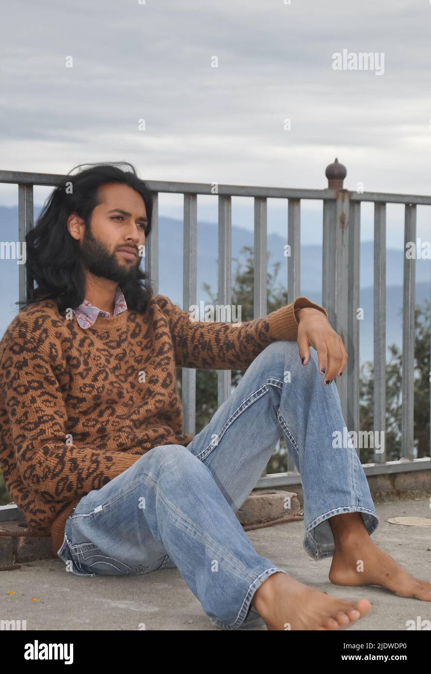 Ein charmanter, langhaariger und bärtiger, junger Mann aus Indien, der zur Seite blickt und mit einer Sicherheitsbarriere vor dem Hintergrund der Berge sitzt Stockfoto