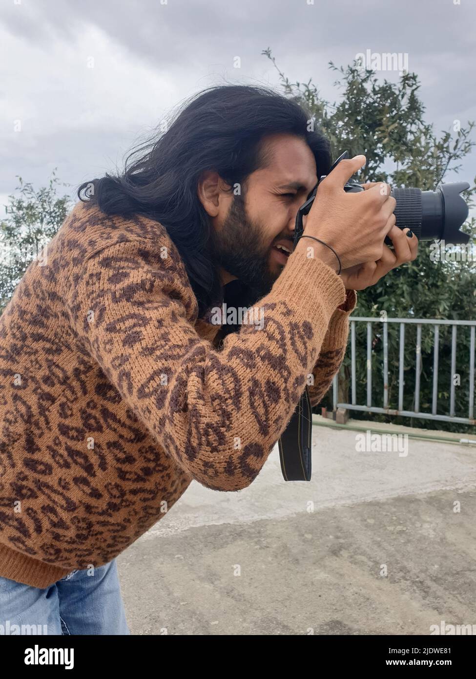 Nahaufnahme eines gut aussehenden jungen Mannes mit langen Haaren und Bart, der mit einer DSLR-Kamera (Digital Single-Lens Reflex) im Freien fotografiert Stockfoto