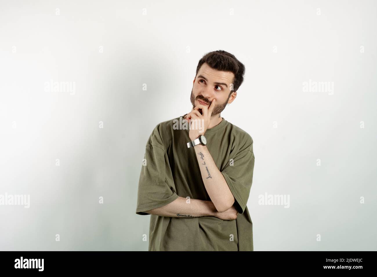 Junger Mann in einem Khaki-T-Shirt, der isoliert auf weißem Hintergrund posiert und in der rechten oberen Ecke die Lippe berührt, denkend. Versuchen Sie, eine Lösung zu finden. Stockfoto