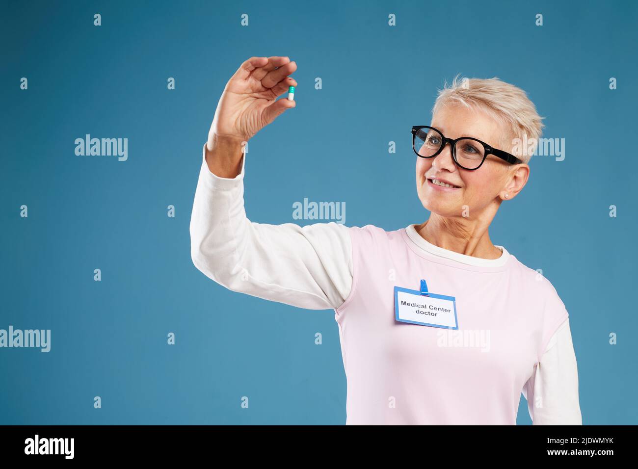 Positive intelligente reife Apothekerin mit Abzeichen der medizinischen Klinik, die auf blauem Hintergrund steht und während der Untersuchung des pi eine Kapsel betrachtet Stockfoto
