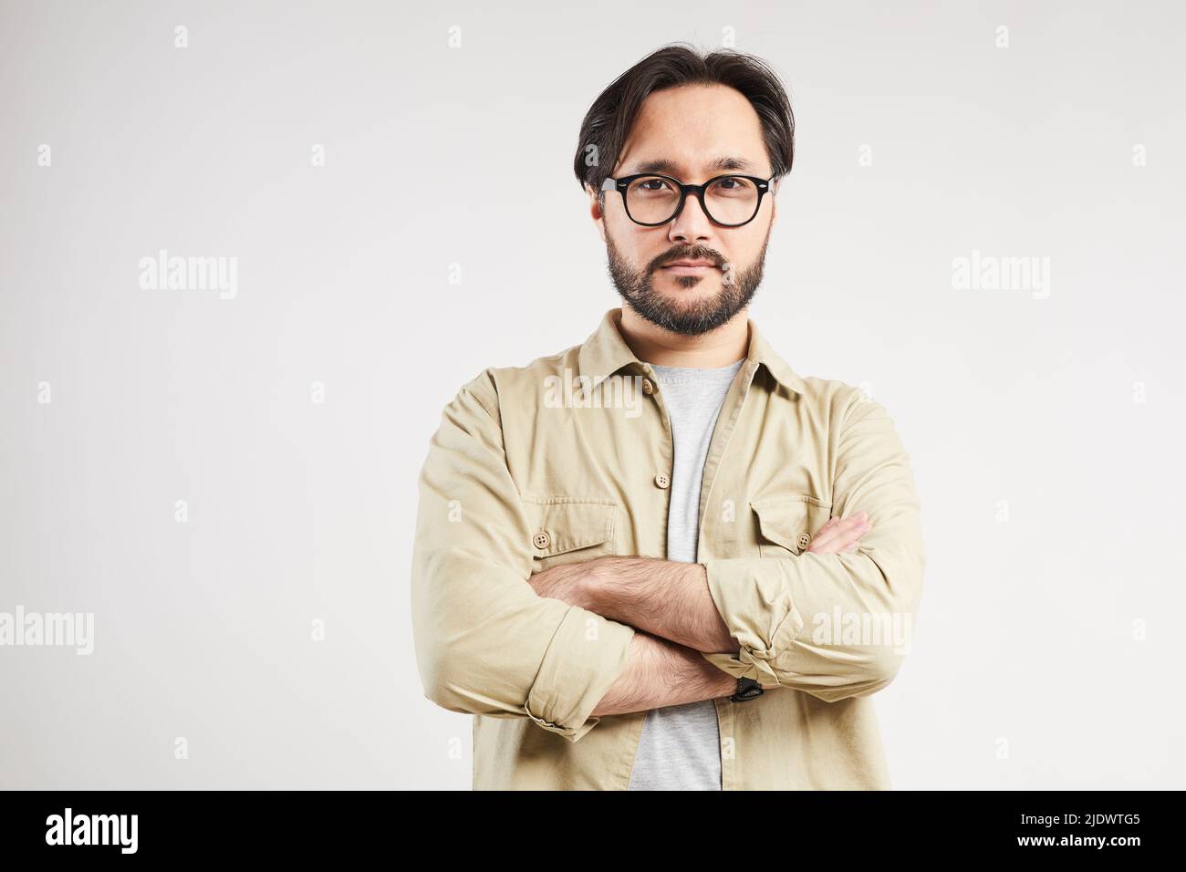 Porträt eines hübschen jungen asiatischen Mannes im Khaki-Hemd mit stilvoller Brille, die die Arme vor grauem Hintergrund kreuzt Stockfoto