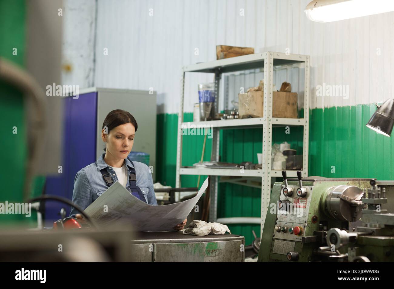 Eine seriöse, konzentrierte Ingenieurin mittleren Alters mit braunen Haaren, die am Schreibtisch stehen und mit Blaupause arbeiten, während sie Details entwirft Stockfoto