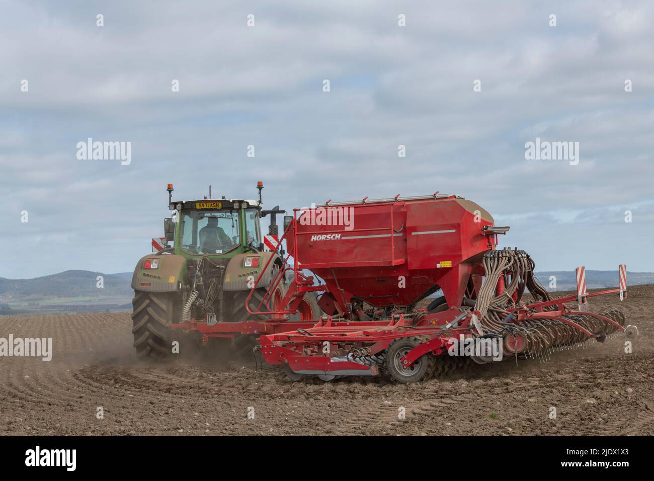 Ein grüner Fendt-Traktor mit einem roten Horsch-Sämaschine, der sich während der Aussaat von Gerste im Frühjahr am Rand eines gepflügten Feldes dreht Stockfoto