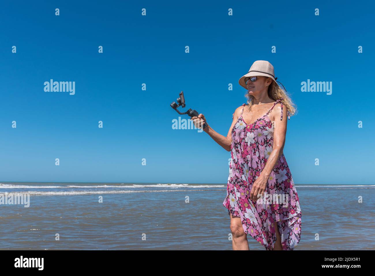 Blonde Frau im Hut und Sommerkleid, die beim Strandspaziergang ein Foto gemacht hat Stockfoto