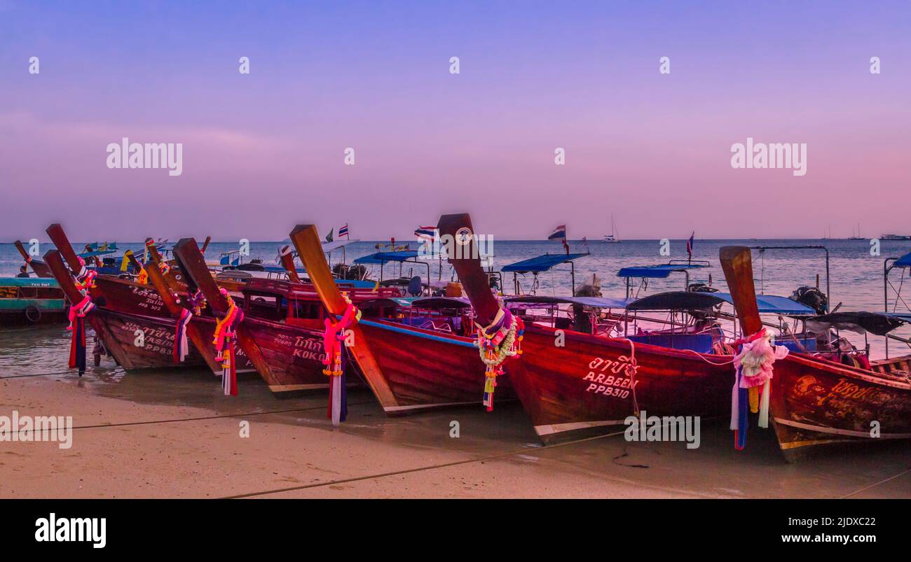 Kho Phi Phi lange Boote standen in einer Reihe am Strand für die Nacht. Sonnenuntergangsfarben am Himmel und am Strand im Vordergrund. Stockfoto