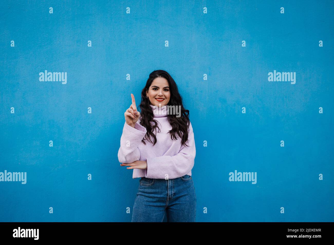 Lächelnde junge, schöne Frau, die die Nummer eins mit Zeigefinger vor der blauen Wand zeigt Stockfoto