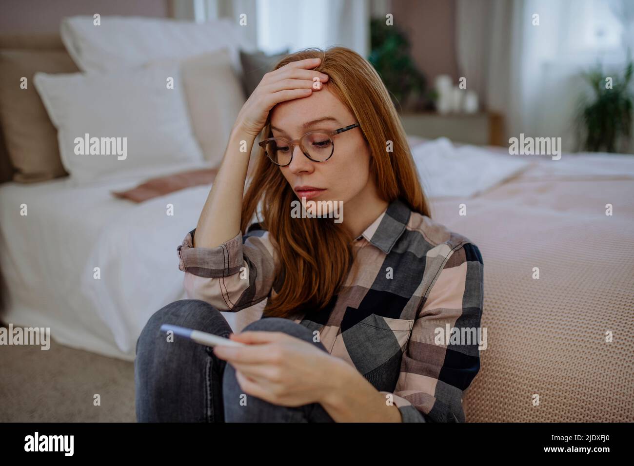 Besorgte Frau mit Schwangerschaftstest-Kit sitzt vor dem Bett zu Hause Stockfoto