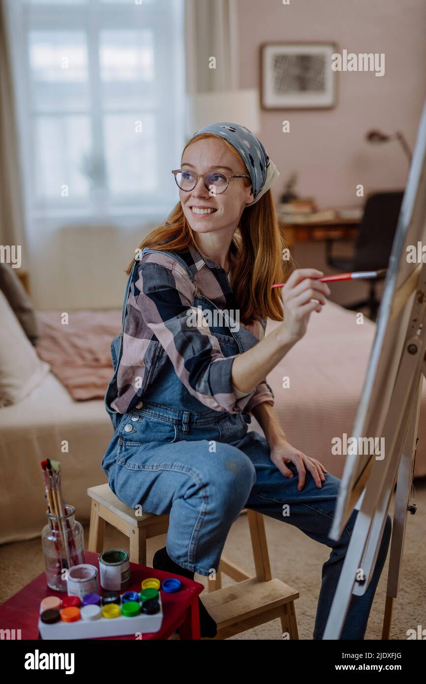 Glückliche Frau mit Pinsel sitzt auf dem Stuhl von der Leinwand des Künstlers zu Hause Stockfoto