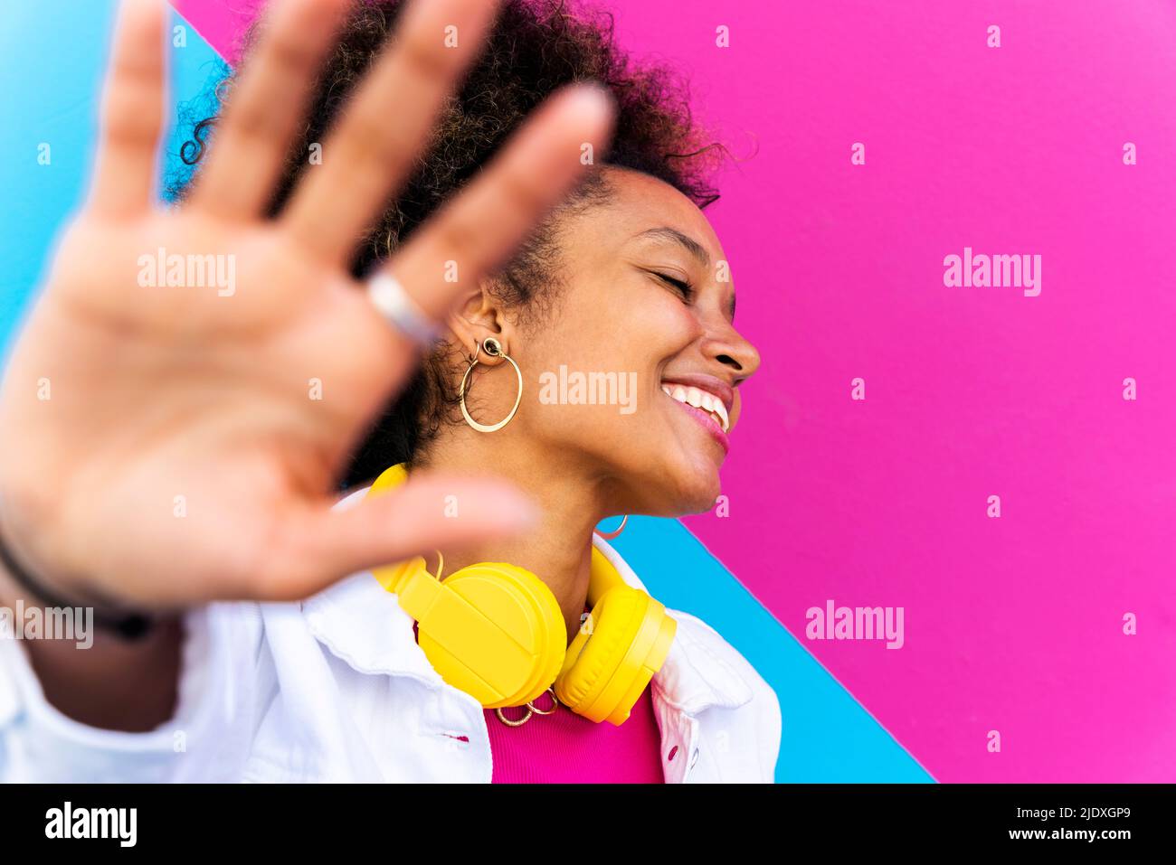 Lächelnde Frau mit geschlossenen Augen und gestikulierenden Stoppschild vor rosa und blauer Wand Stockfoto