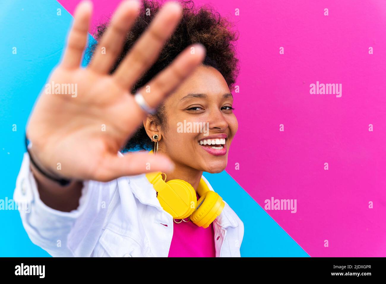 Fröhliche junge Frau mit kabellosen Kopfhörern, die das Stoppschild vor einer rosa und blauen Wand staufelt Stockfoto