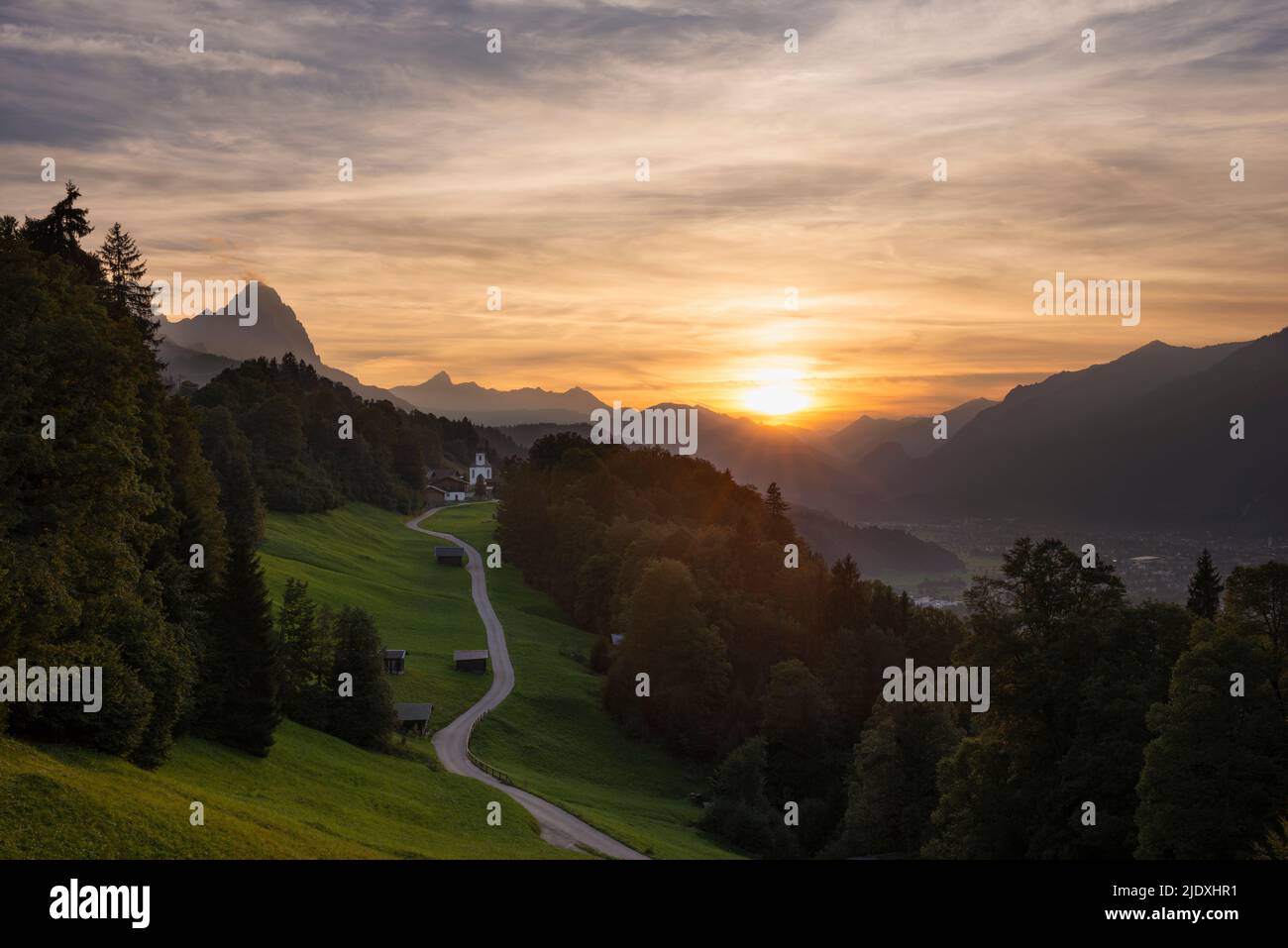 Deutschland, Bayern, Wamberg, Sonne untergeht über eine kleine Straße, die zu einem abgelegenen Dorf in den Wettersteinbergen führt Stockfoto