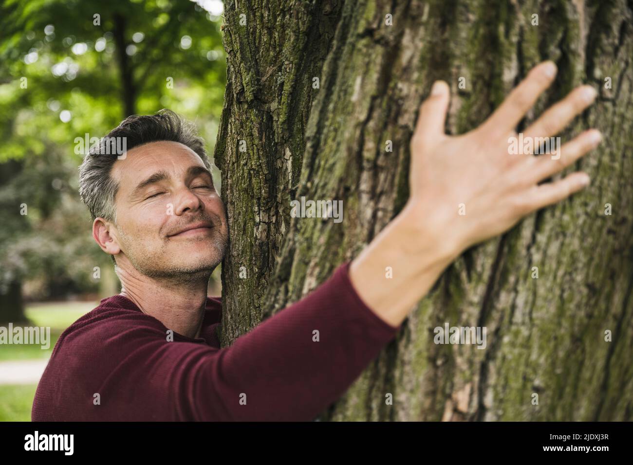 Lächelnder Mann mit geschlossenen Augen umarmter Baum im Park Stockfoto