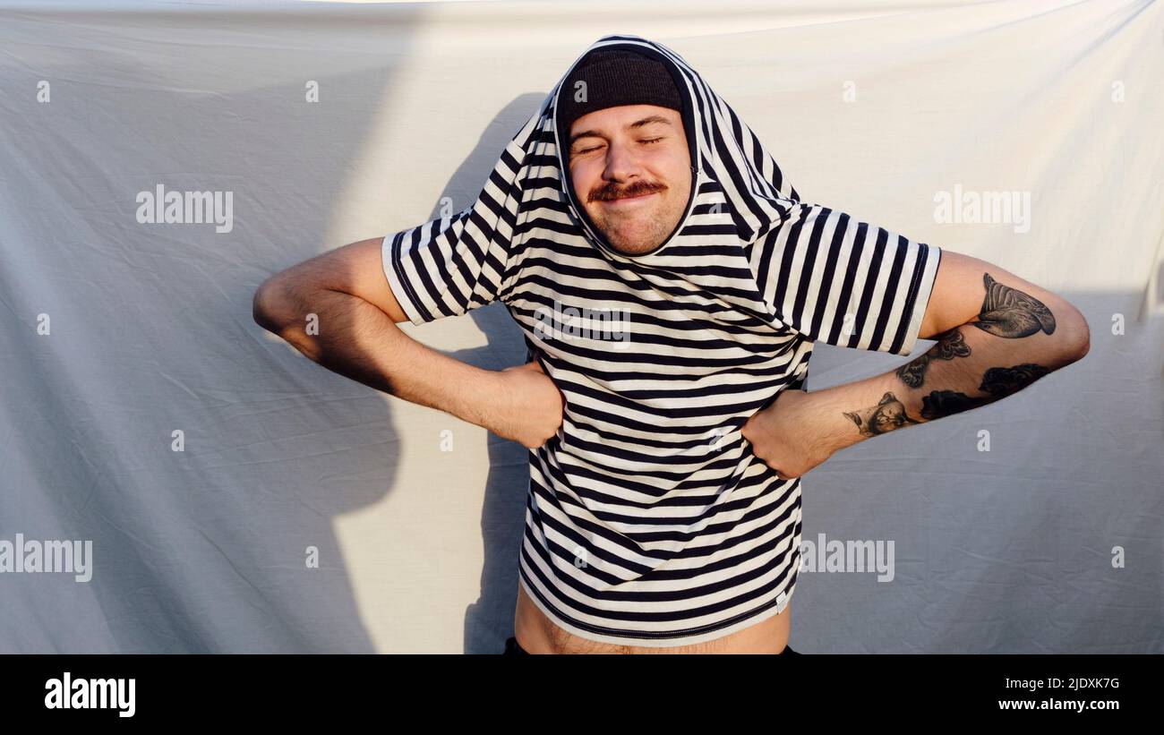 Lächelnder Mann in gestreiftem T-Shirt mit Akimbo-Armen vor weißem Hintergrund Stockfoto