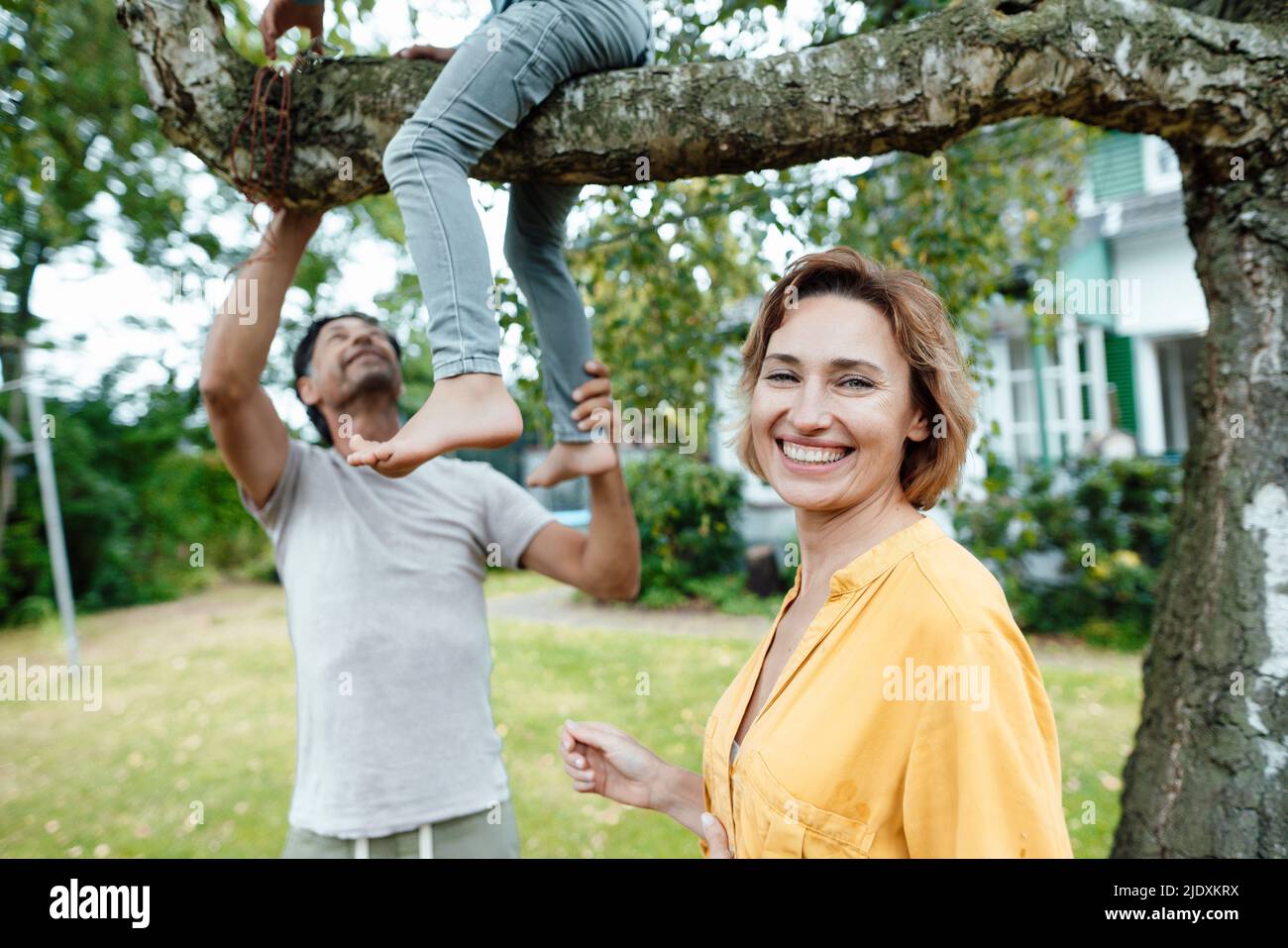 Glückliche Frau, die vor dem Mann steht und den Sohn anschaut, der auf dem Baum im Hinterhof sitzt Stockfoto
