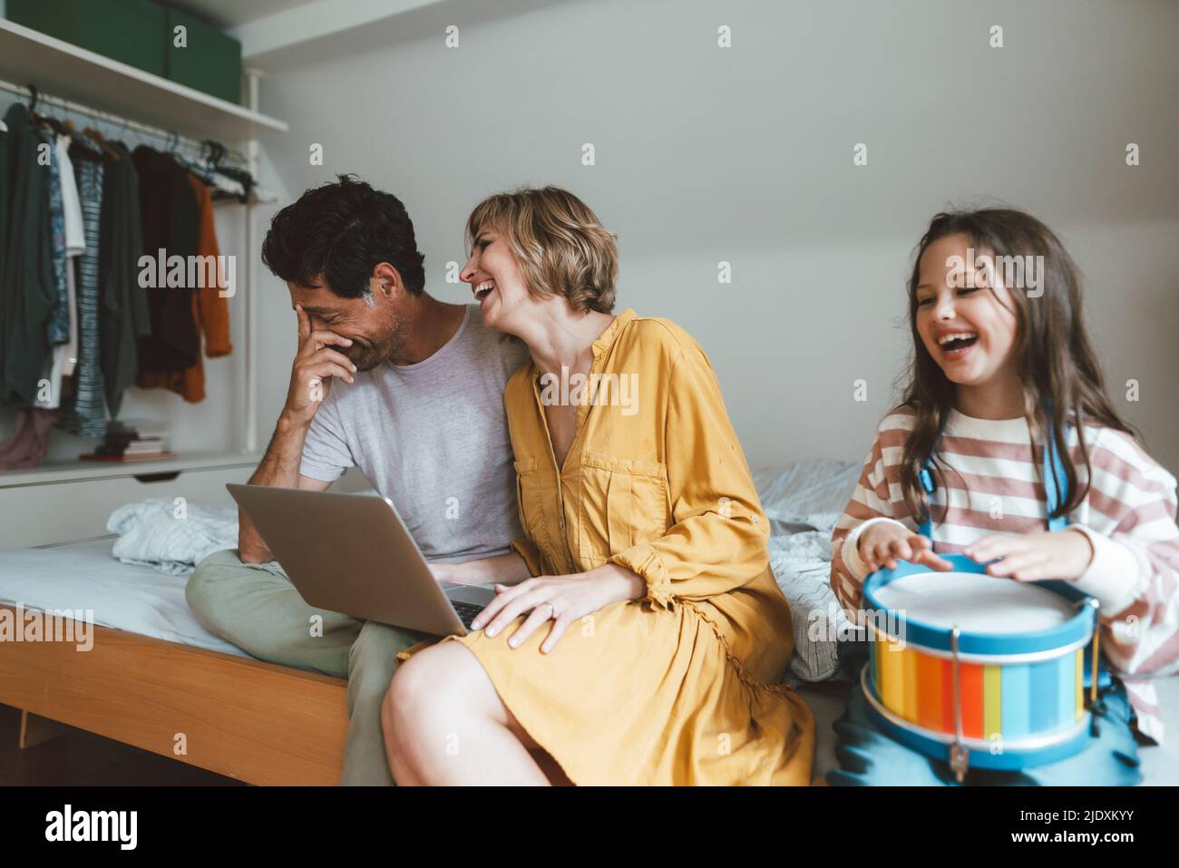 Glücklicher Mann und Frau sitzen mit Laptop von Tochter spielen Spielzeug Trommel auf dem Bett zu Hause Stockfoto