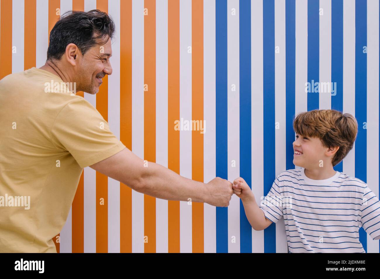 Lächelnder Mann, der dem Sohn eine Faust vor einer orange und blau gestreiften Wand gab Stockfoto