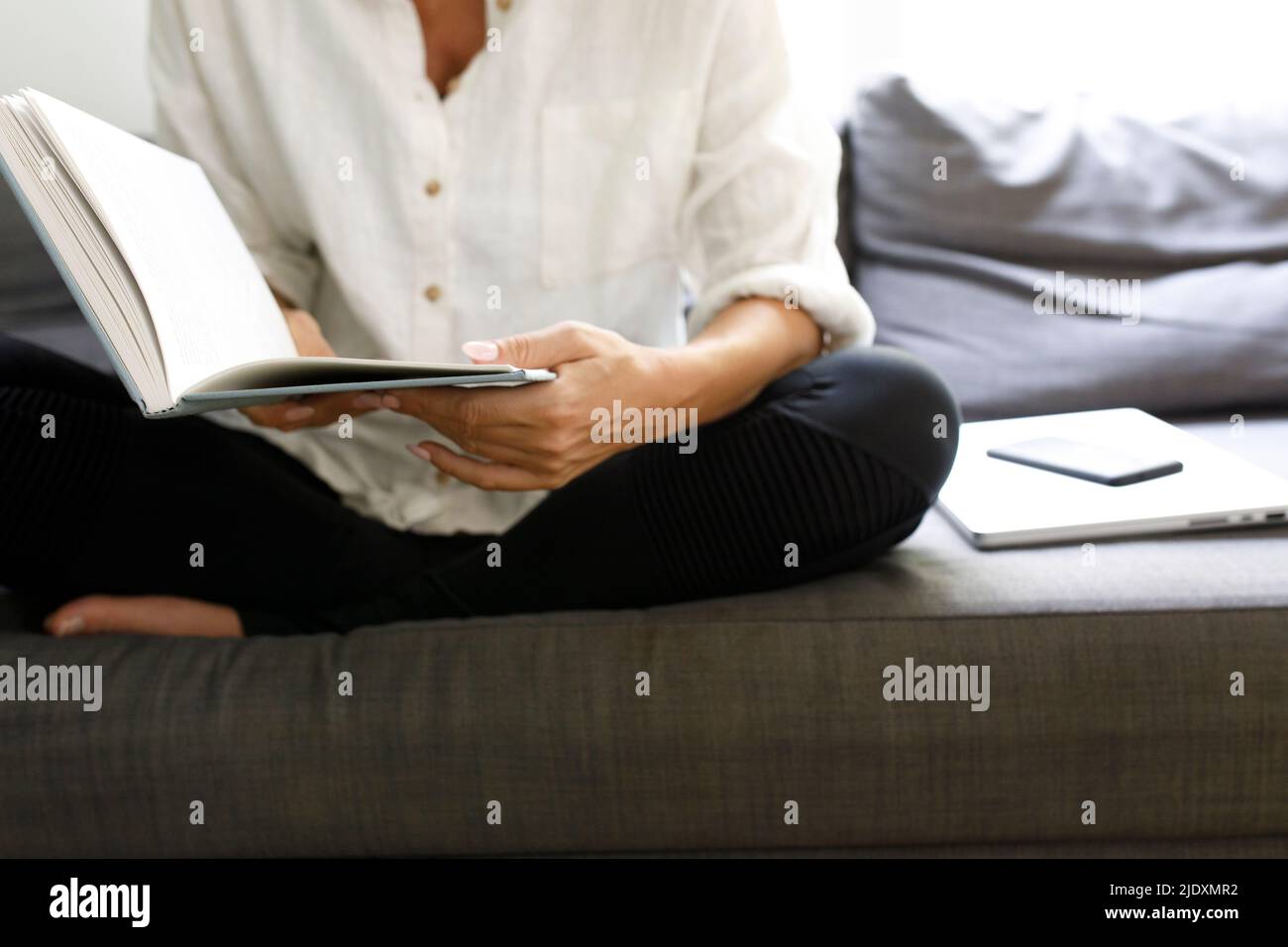 Frau, die Buch liest, sitzt mit drahtlosen Technologien auf dem Sofa zu Hause Stockfoto