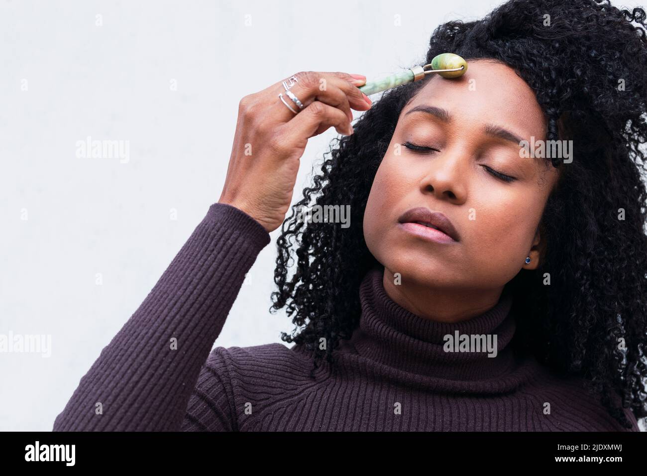 Frau mit lockigen Haaren mit Jaderolle auf dem Gesicht vor weißem Hintergrund Stockfoto