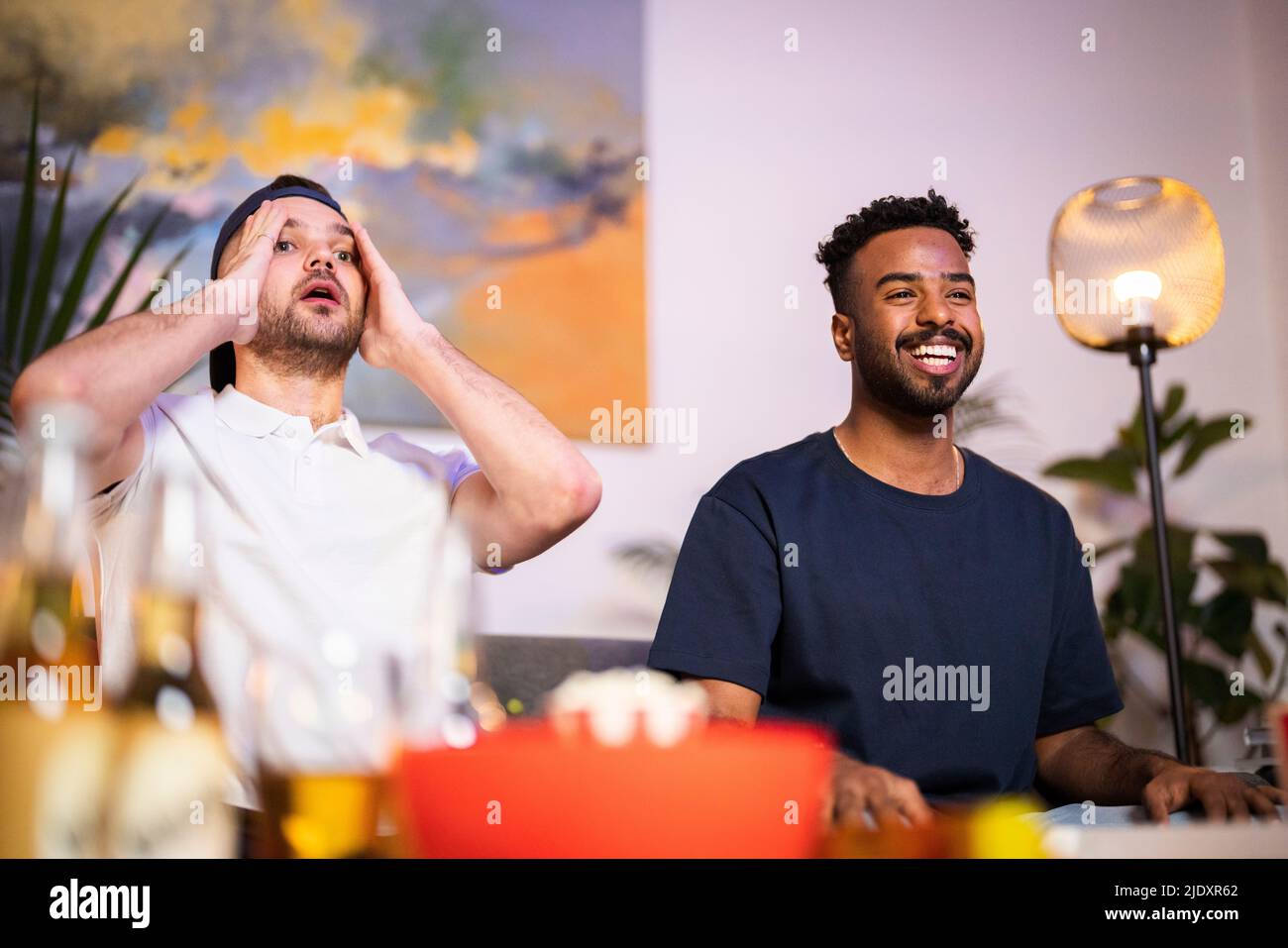 Fröhlicher junger Mann, der von einem enttäuschten Freund sitzt und sich zu Hause ein Fußballspiel ansieht Stockfoto