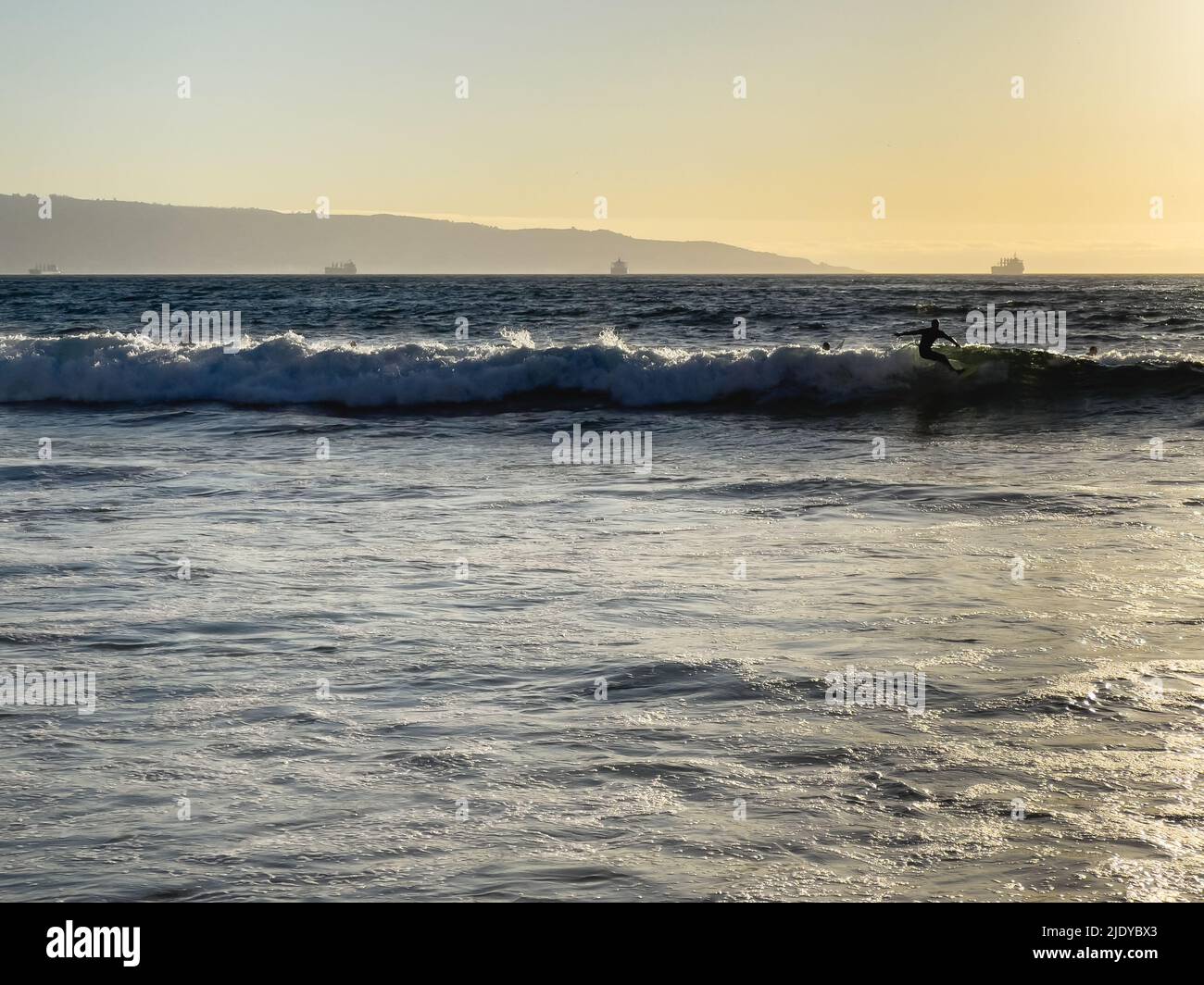 Mann Silhouette Surfen Wellen bei Sonnenuntergang am Strand mit Frachtschiffen durch Valparaiso Hafen im Hintergrund in Chile. Sommerkonzept für Wassersport Stockfoto