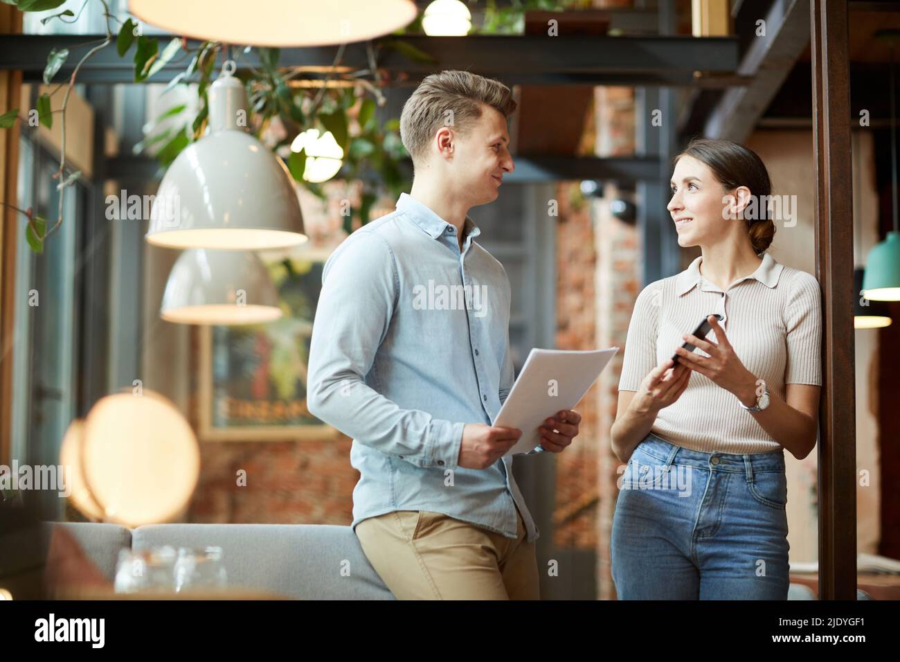 Positiv zufriedene junge Kollegen in lässiger Kleidung stehen im gemütlichen Café und nutzen Smartphone- und Marktanalyse-Bericht während der Online-Ausarbeitung Stockfoto