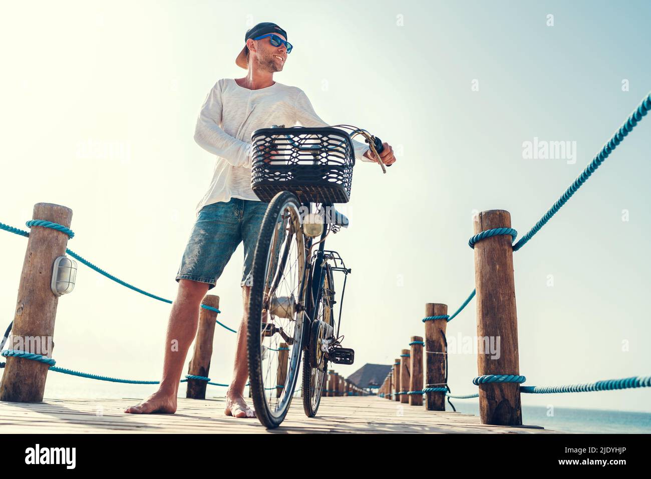 Porträt eines glücklichen lächelnden barfuss-Mannes, gekleidet in leichte Sommerkleidung und Sonnenbrille, der auf dem hölzernen Seebrücke ein Fahrrad fährt. Sorgloser Urlaub in Stockfoto