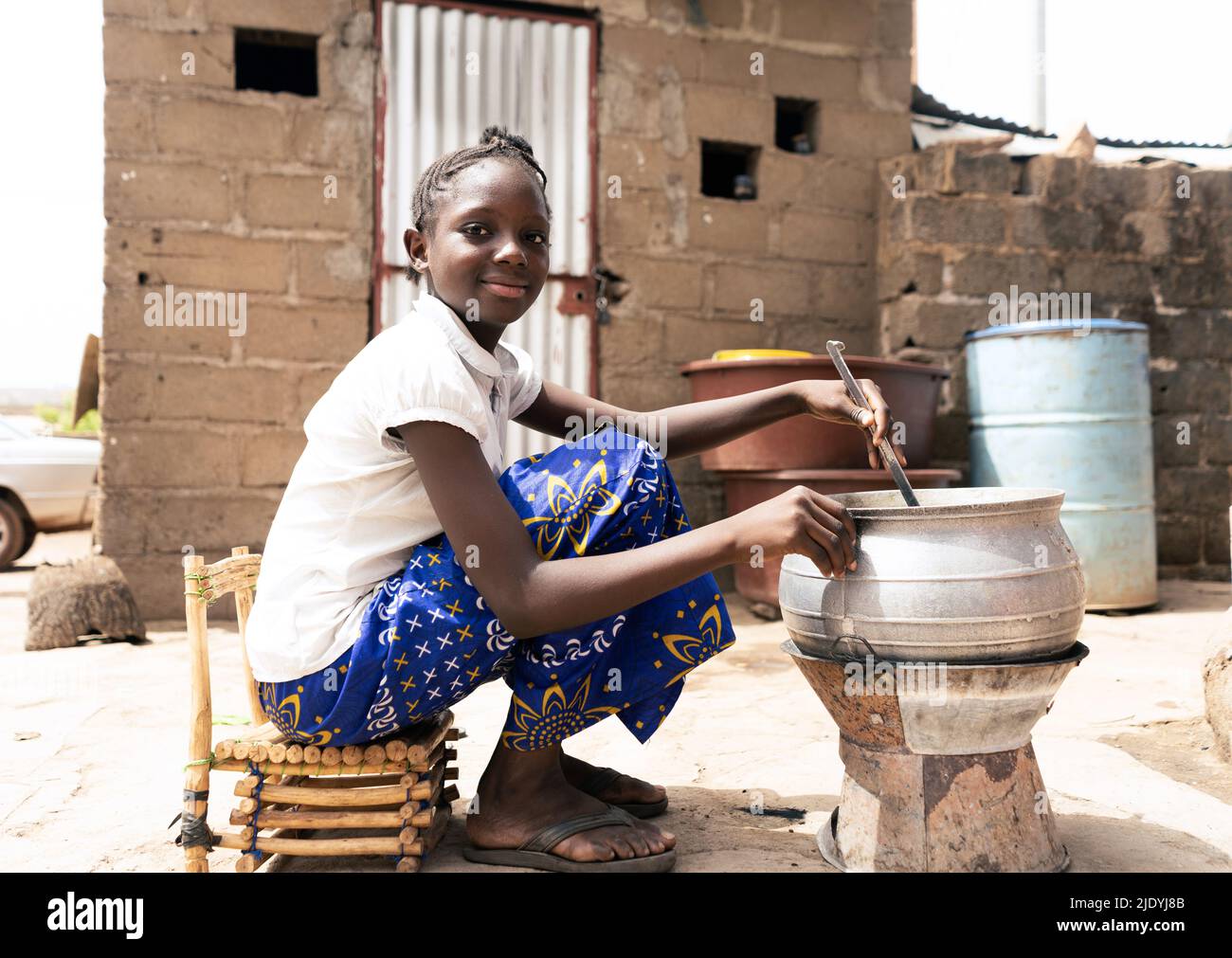 Nettes kleines afrikanisches Mädchen, das vor einem Ofen sitzt, um in einer Großstadt wie Bamako in Mali Essen für ihre Familie zu kochen Stockfoto