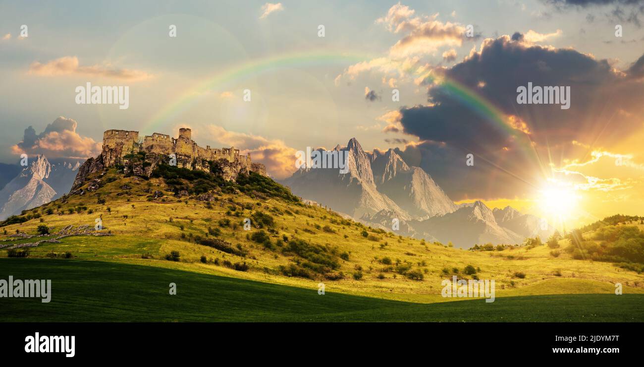 Schloss auf dem Hügel unter einem Regenbogen bei Sonnenuntergang. Composite Fantasy-Landschaft. Wiese im Vordergrund. Felsige Gipfel des Grats in der Ferne Stockfoto