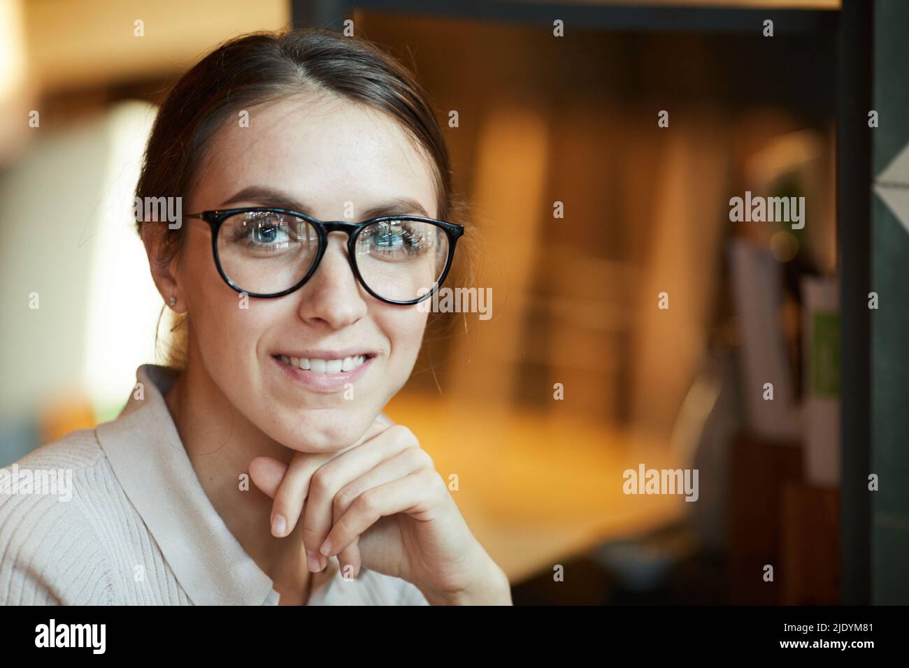 Positive junge hübsche Dame mit freundischem Lächeln, die eine stilvolle Brille trägt, die den Kopf an die Hand lehnt und die Kamera anschaut Stockfoto