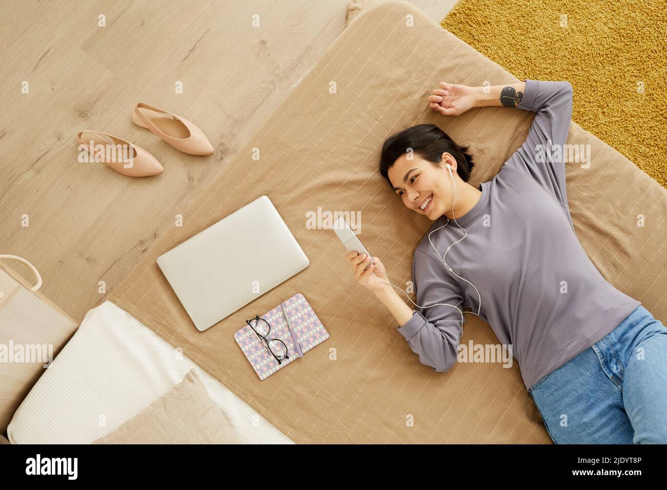 Direkt über dem Bild einer glücklichen jungen Frau mit kurzen Haaren, die auf dem Bett liegt und die Online-Kommunikation mit einem Freund über die mobile App genießt Stockfoto