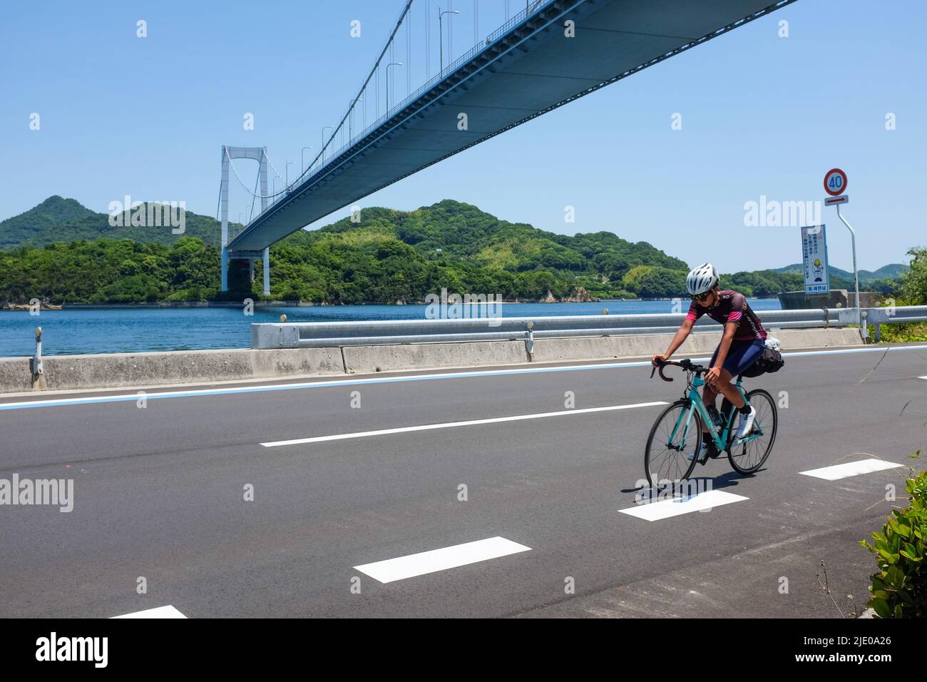 Ein Radfahrer, der auf der Shimanami Kaido-Radroute zwischen Onomichi und Imabari in Japan mit der Akinada-Brücke im Hintergrund unterwegs ist. Stockfoto