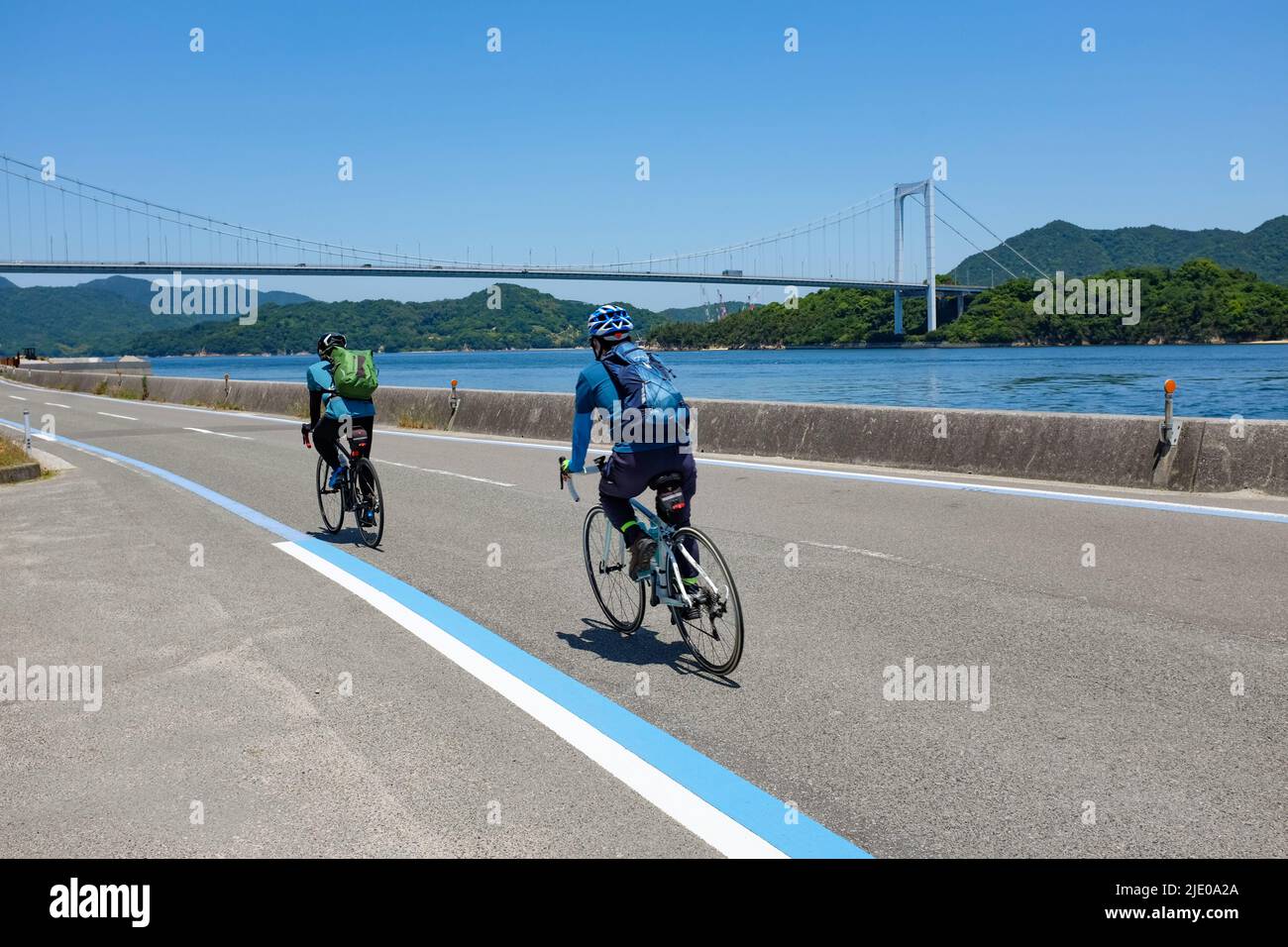 Radfahrer fahren entlang der Shimanami Kaido-Radroute zwischen Onomichi und Imabari in Japan, mit der Akinada-Brücke im Hintergrund. Stockfoto