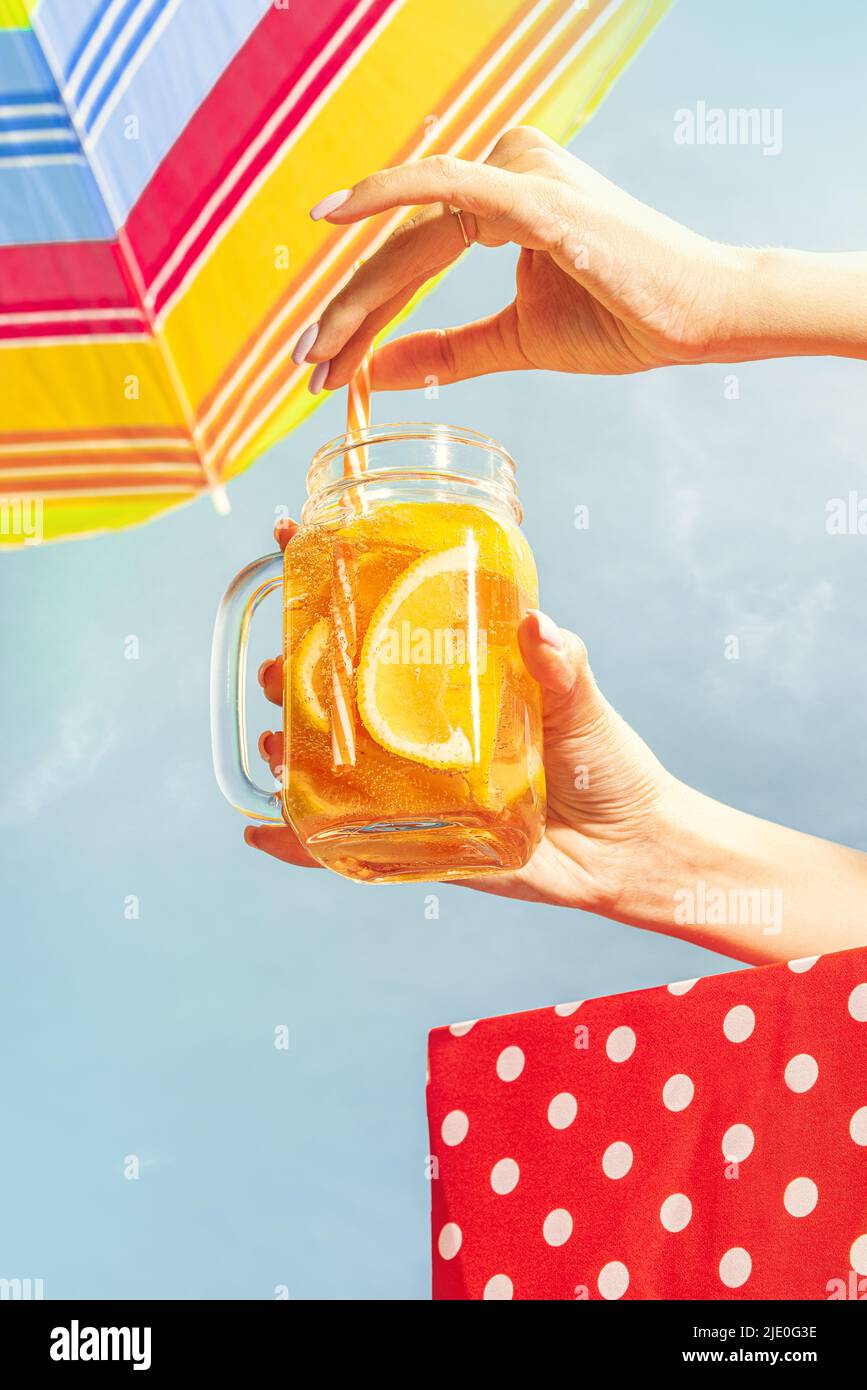 Weibliche Hand hält Glaskrug mit frostiger oranger Limonade, Cocktail auf blauem sonnigen Himmel Hintergrund. Urlaub, Glück, Sommerstimmung. Retro-Style Stockfoto