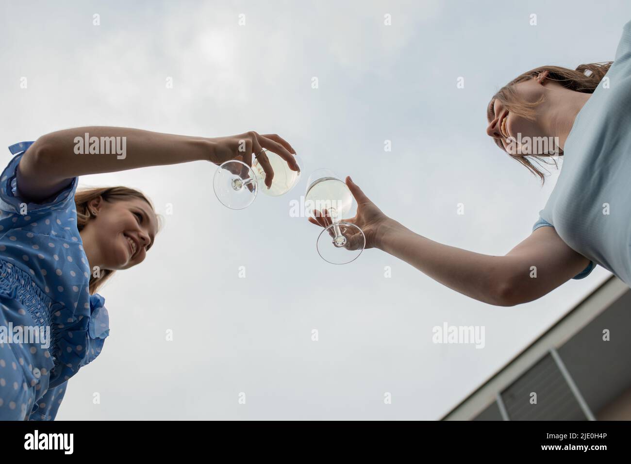 Glückliche junge Mädchen stehen einander gegenüber, klirren Gläser und trinken Wein aus der Nähe von unten, Himmel Hintergrund. Spaß haben und genießen Stockfoto