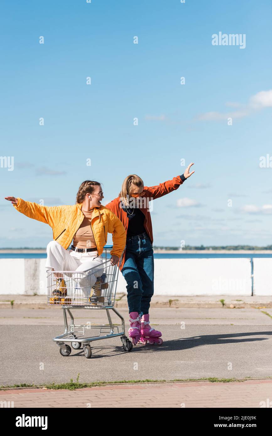 Junge Frau, die im Einkaufswagen in der Nähe von Freunden sitzt, die im Freien Rollschuhe reiten Stockfoto