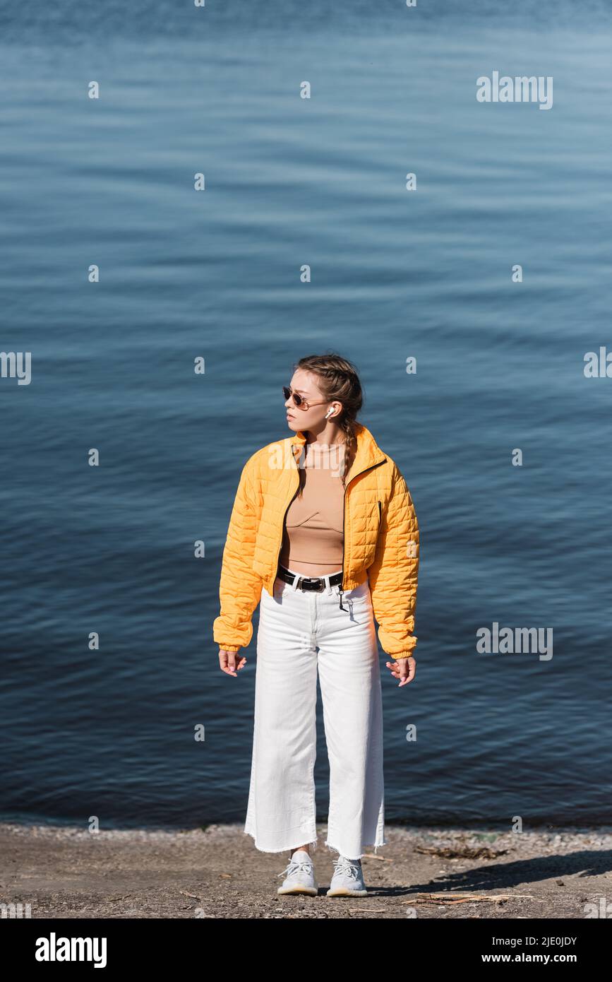 Frau in voller Länge in weißer Hose und gelber Jacke mit Blick auf das Flussufer Stockfoto