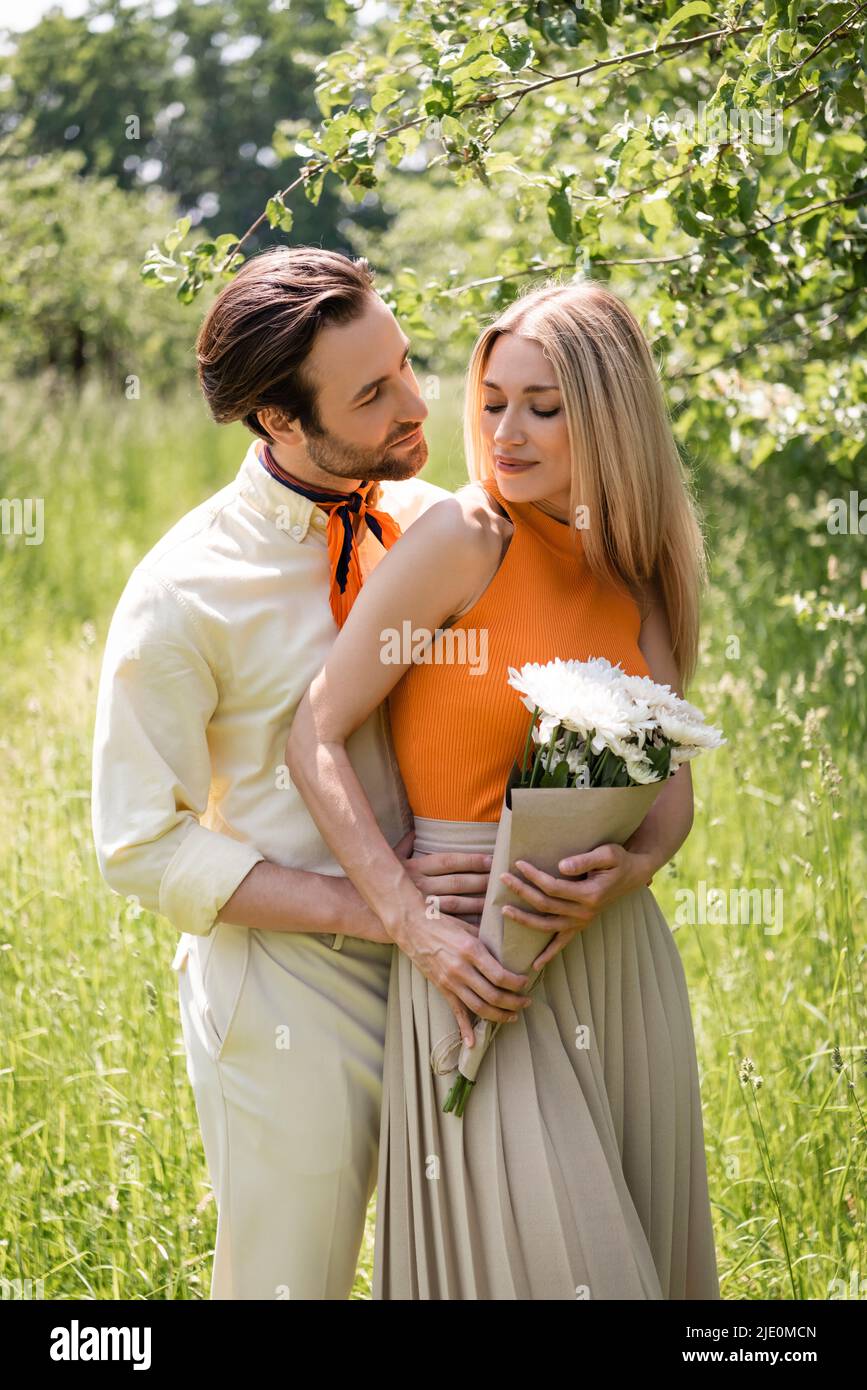 Junger Mann umarmt lächelnde Freundin mit Blumenstrauß im Sommerpark Stockfoto