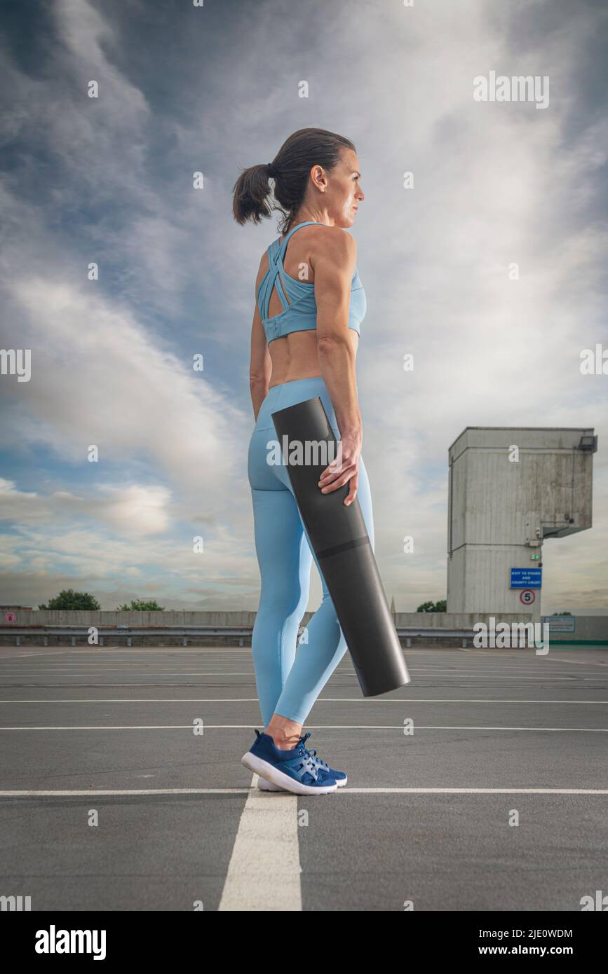Sportliche Frau, die eine aufgerollte Yoga-Matte trägt, urbaner Hintergrund. Stockfoto