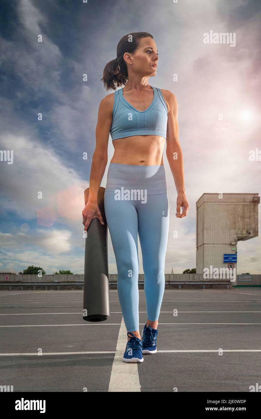 Sportliche Frau, die eine aufgerollte Yoga-Matte trägt, urbaner Hintergrund. Stockfoto