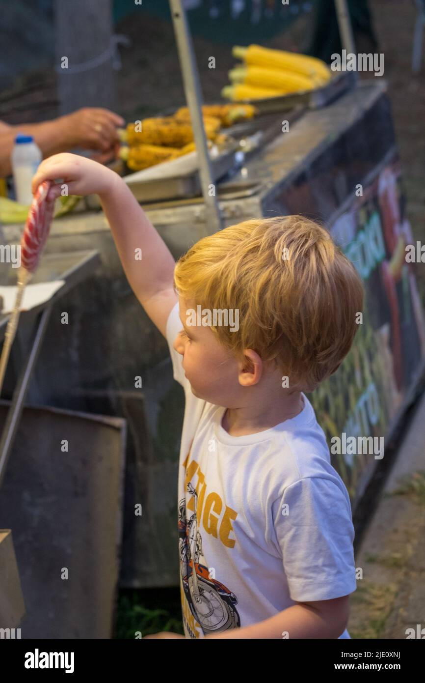 Netter kleiner Junge, der während eines Festivals oder einer Veranstaltung einen Lutscher von einem Maisstandplatz nimmt Stockfoto