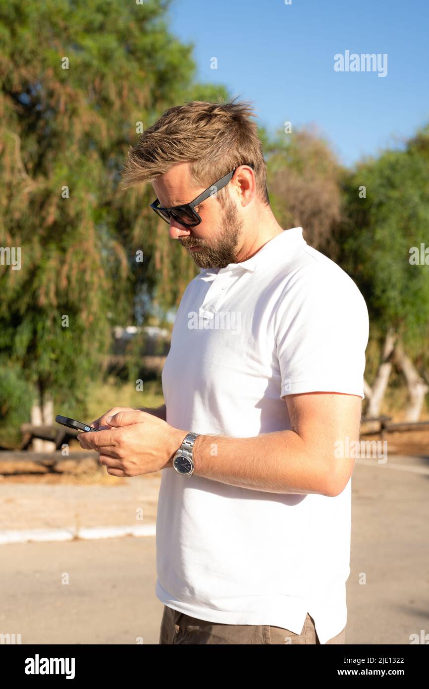 Außenaufnahme eines schönen bärtigen jungen Mannes in Sonnenbrillen, der an warmen sonnigen Tagen über dem Stadtgarten posiert und mit einem Smartphone ein weißes Poloshirt trägt. Stockfoto