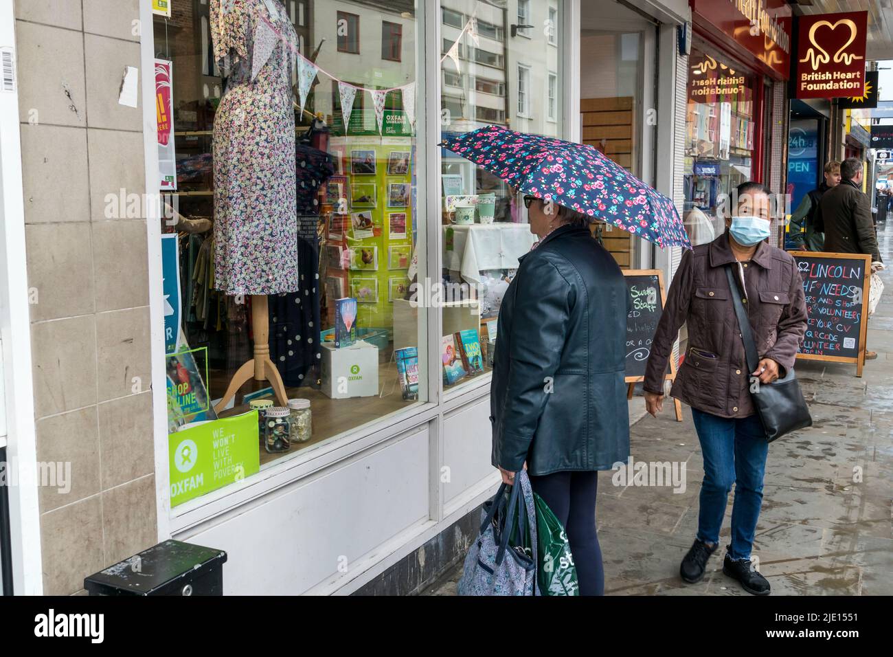 Dame mit Regenschirm, die das Kleid im OXFAM Schaufenster der High Street Lincoln City 2022 anschaut Stockfoto