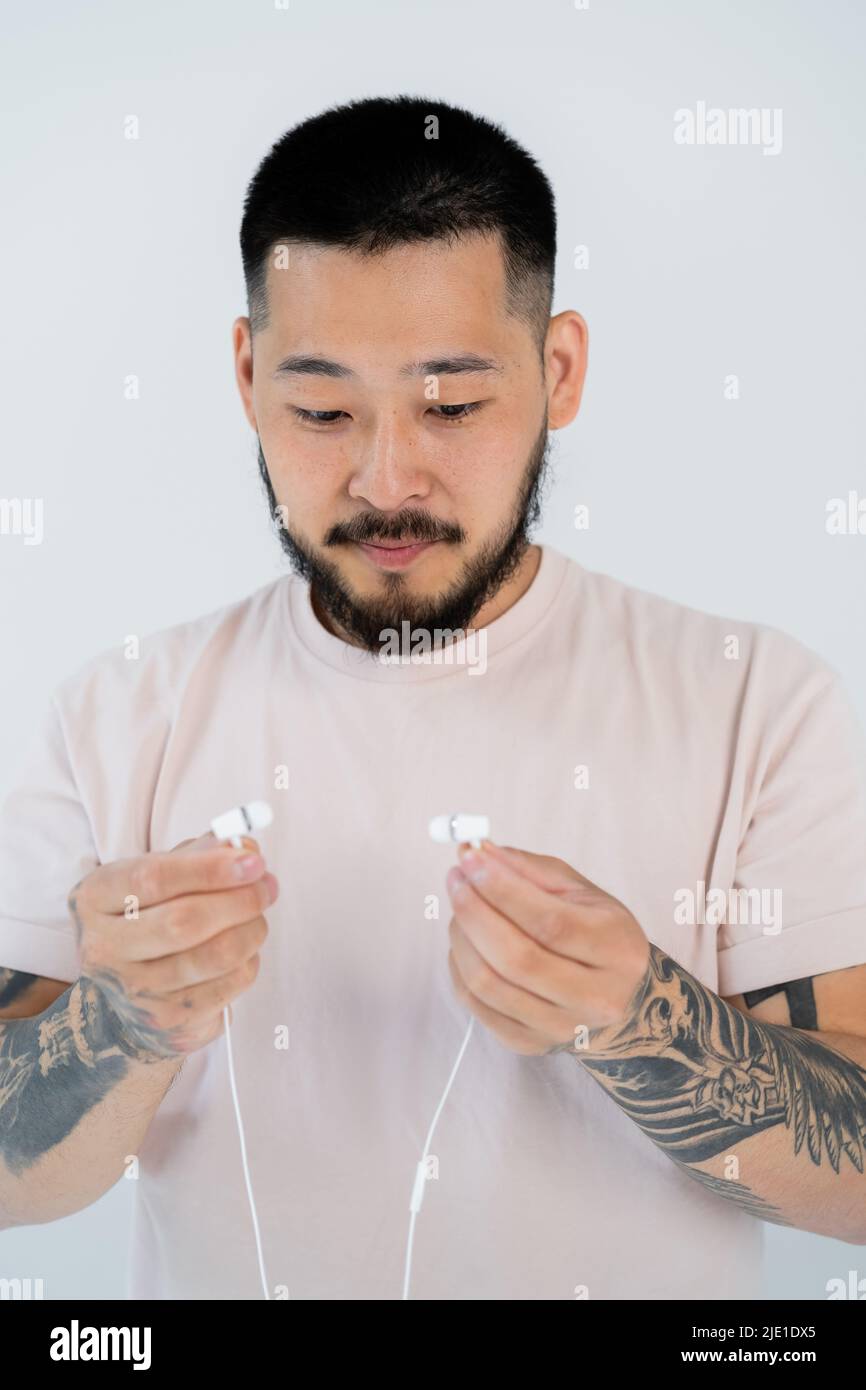 Porträt eines tätowierten asiatischen Mannes in T-Shirt, der auf grau isolierte kabelgebundene Ohrhörer blickt Stockfoto
