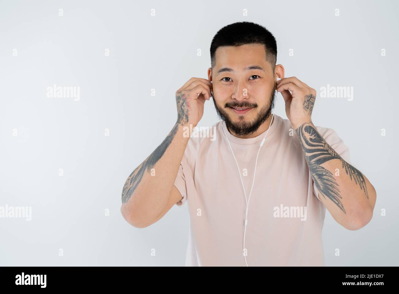 Tätowierter asiatischer Mann im T-Shirt lächelt, während er kabelgebundene Kopfhörer trägt und die Kamera isoliert auf Grau anschaut Stockfoto