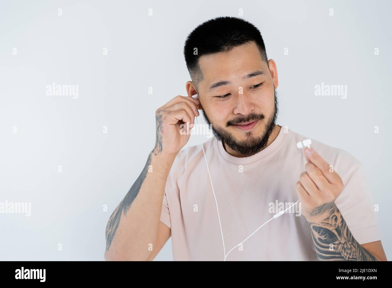 Tätowierte asiatische Mann in T-Shirt lächelnd und tragen Kabel-Kopfhörer isoliert auf grau Stockfoto