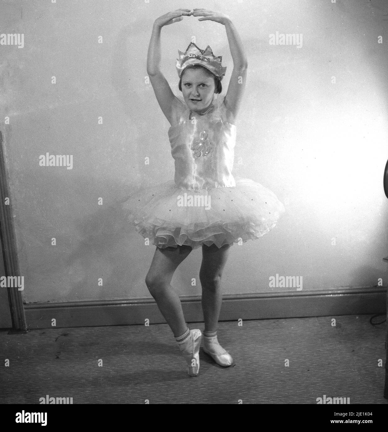 1950s, historisch, ein kleines Mädchen in ihrem Ballettkostüm, spitzenschuhe und eine Krone auf ihrem Kopf, eine Pose in Innenräumen, Stockport, England, Großbritannien. Das Ballettkostüm, ein Rüschenrock, der an einem schnittig anliegenden Oberteil befestigt ist, wird auch als Tutu bezeichnet. Stockfoto